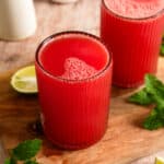 Refreshing homemade watermelon juice served in two ribbed glasses on a wooden board with fresh mint leaves and lime wedges, featuring a vibrant red color and naturally frothy top.