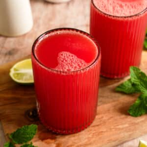 Refreshing homemade watermelon juice served in two ribbed glasses on a wooden board with fresh mint leaves and lime wedges, featuring a vibrant red color and naturally frothy top.