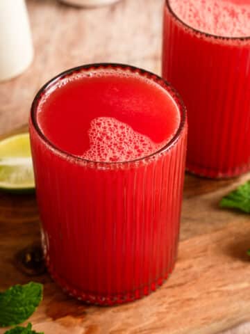 Refreshing homemade watermelon juice served in two ribbed glasses on a wooden board with fresh mint leaves and lime wedges, featuring a vibrant red color and naturally frothy top.