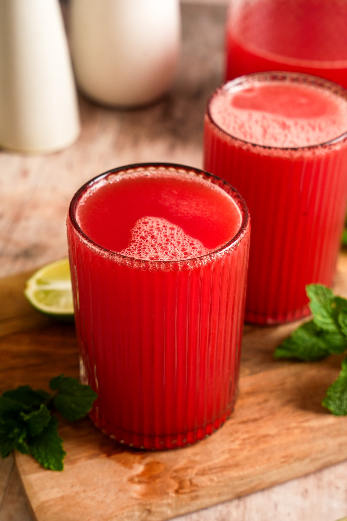 Fresh watermelon juice served in two ribbed glasses on a wooden board, with lime wedges, fresh mint, and a pitcher of juice in the background.