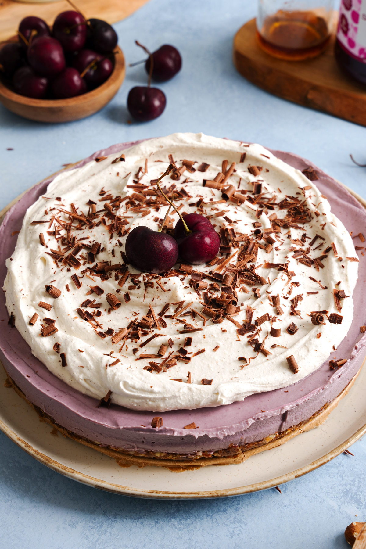 Raw vegan cherry almond cheesecake topped with fluffy dairy-free whipped cream, fresh cherries, and dark chocolate shavings on a date and almond crust, served on a white plate.
