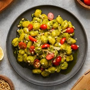 Close-up of homemade vegan pesto gnocchi coated in a vibrant basil pesto and served on a black plate. The dish is topped with juicy cherry tomatoes, toasted pine nuts, and shaved vegan Parmesan, creating a fresh, colorful, and comforting plant-based meal.