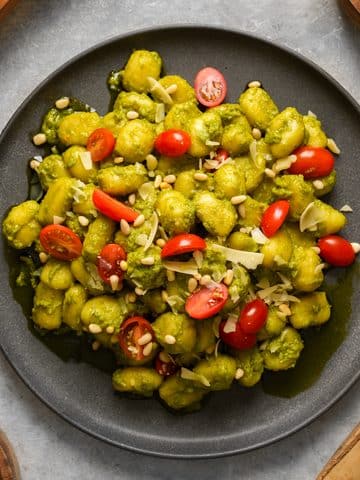 Close-up of homemade vegan pesto gnocchi coated in a vibrant basil pesto and served on a black plate. The dish is topped with juicy cherry tomatoes, toasted pine nuts, and shaved vegan Parmesan, creating a fresh, colorful, and comforting plant-based meal.