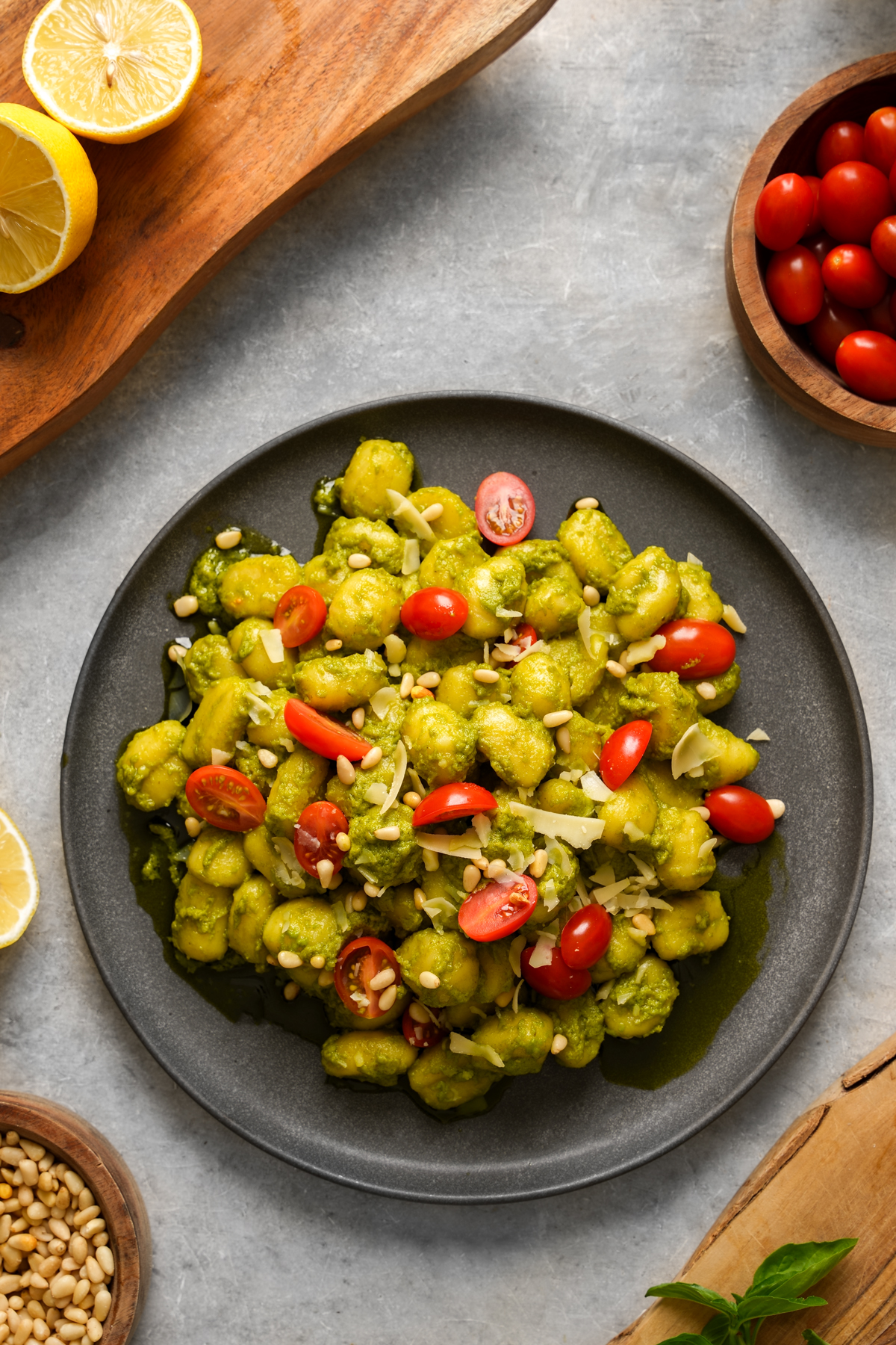 A black plate filled with vegan pesto gnocchi topped with juicy cherry tomatoes, toasted pine nuts, and shaved vegan Parmesan. The dish is garnished with fresh basil and surrounded by lemons, cherry tomatoes, pine nuts, and basil on a light gray surface, creating a bright Mediterranean-inspired presentation.