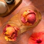 Overhead view of thick and creamy Ninja Creami tropical sorbet with vibrant mango and dragon fruit swirls scooped into two glasses, served beside a red hibiscus flower.
