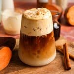 Close-up of a layered iced sweet potato maple latte with creamy cinnamon cold foam in a ribbed glass. The fall-inspired vegan coffee drink is served with sweet potatoes, maple syrup, oat milk, and cinnamon sticks on a rustic wooden board.