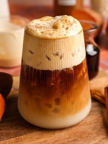 Close-up of a layered iced sweet potato maple latte with creamy cinnamon cold foam in a ribbed glass. The fall-inspired vegan coffee drink is served with sweet potatoes, maple syrup, oat milk, and cinnamon sticks on a rustic wooden board.