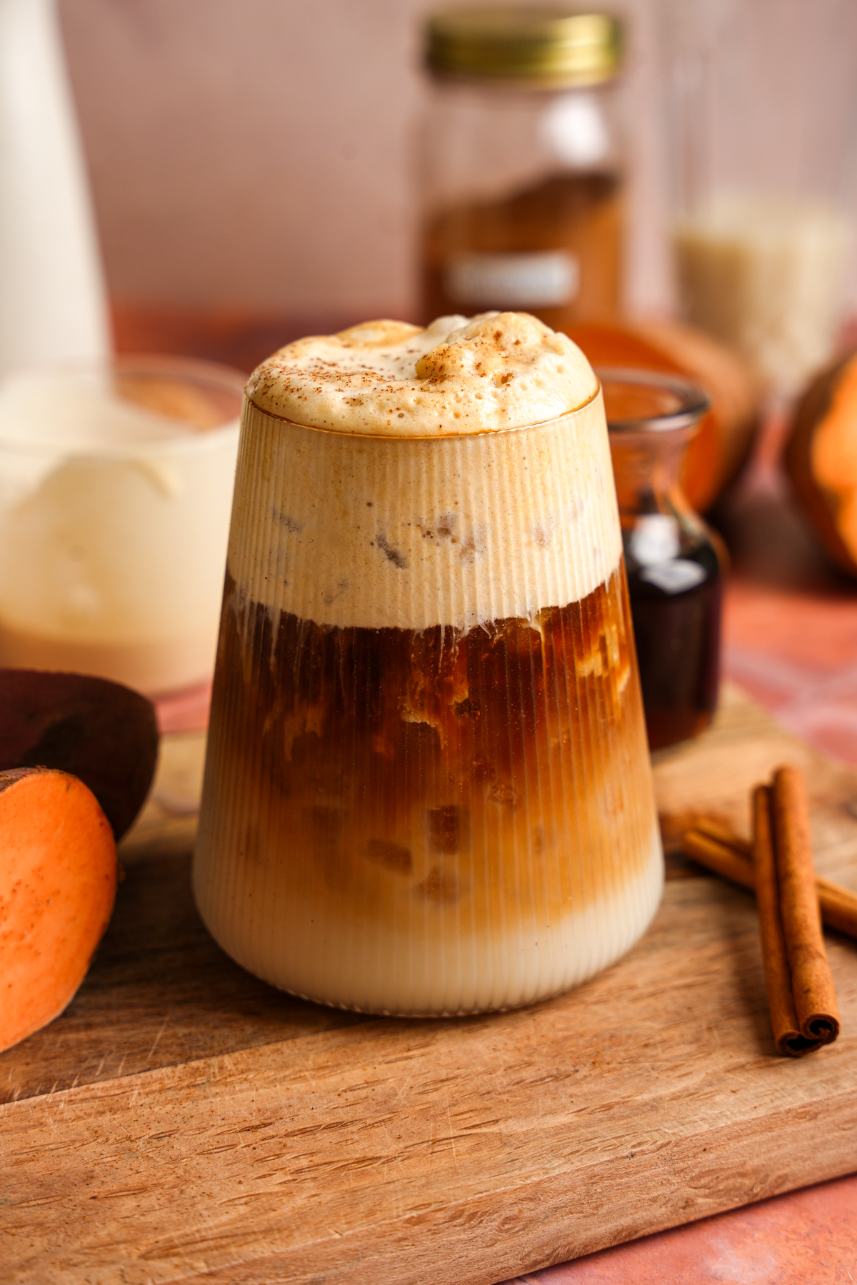 A layered iced sweet potato maple latte in a ribbed glass topped with creamy cinnamon cold foam. The fall-inspired coffee drink is garnished with cinnamon and photographed on a wooden board with sweet potatoes, maple syrup, oat milk, and cinnamon sticks in the background.